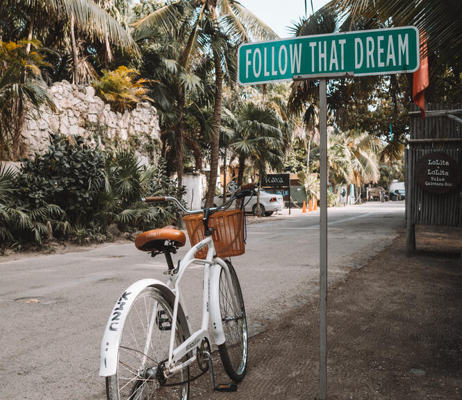 A bicycle on a tropical street by a sign which reads "Follow that dream"